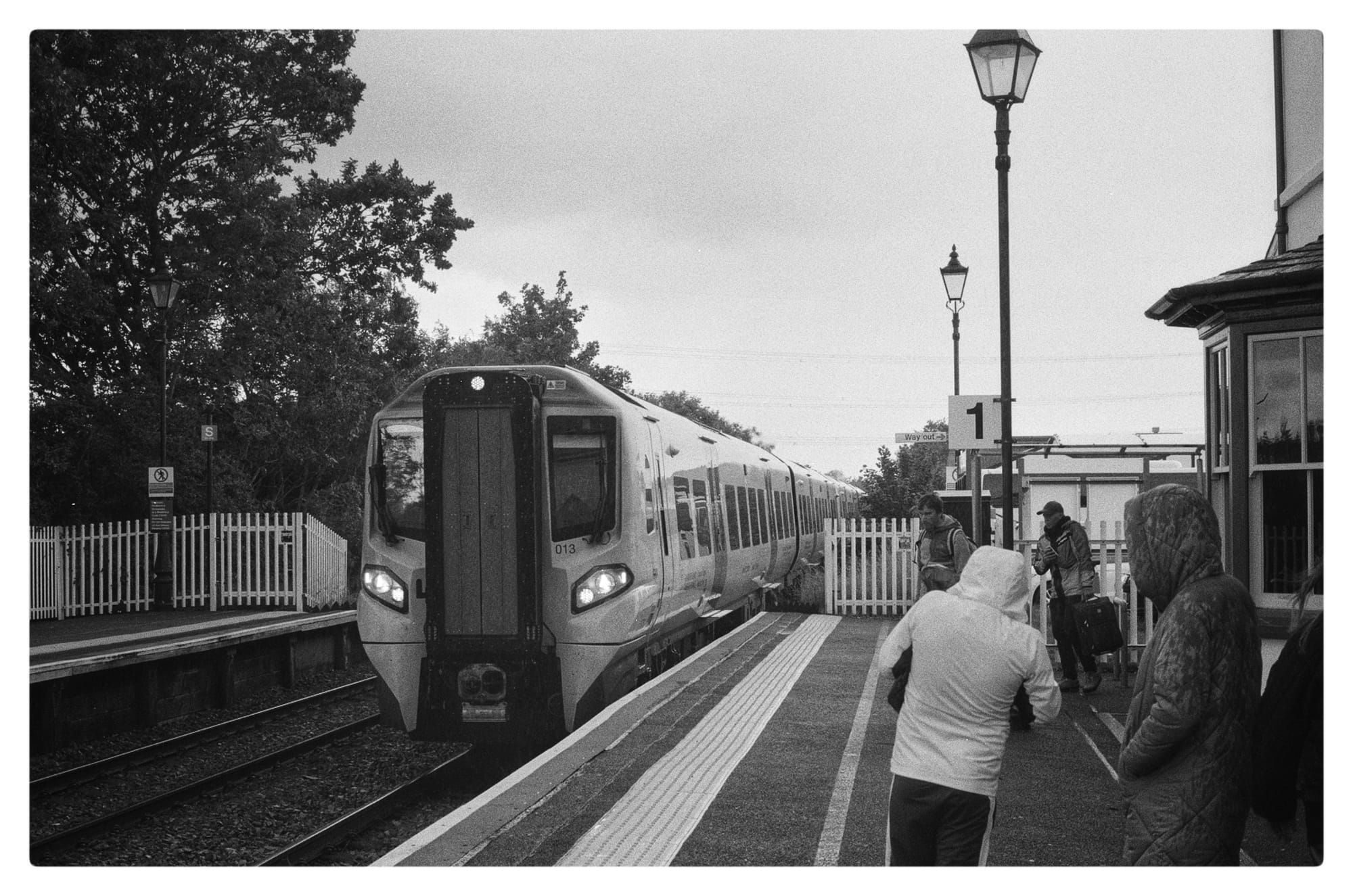 Black and white film photo of Llanfairpwllgwyngyllgogerychwyrndrobwllllantysiliogogogoch train station with a train coming in with a group of people waiting to enter it.